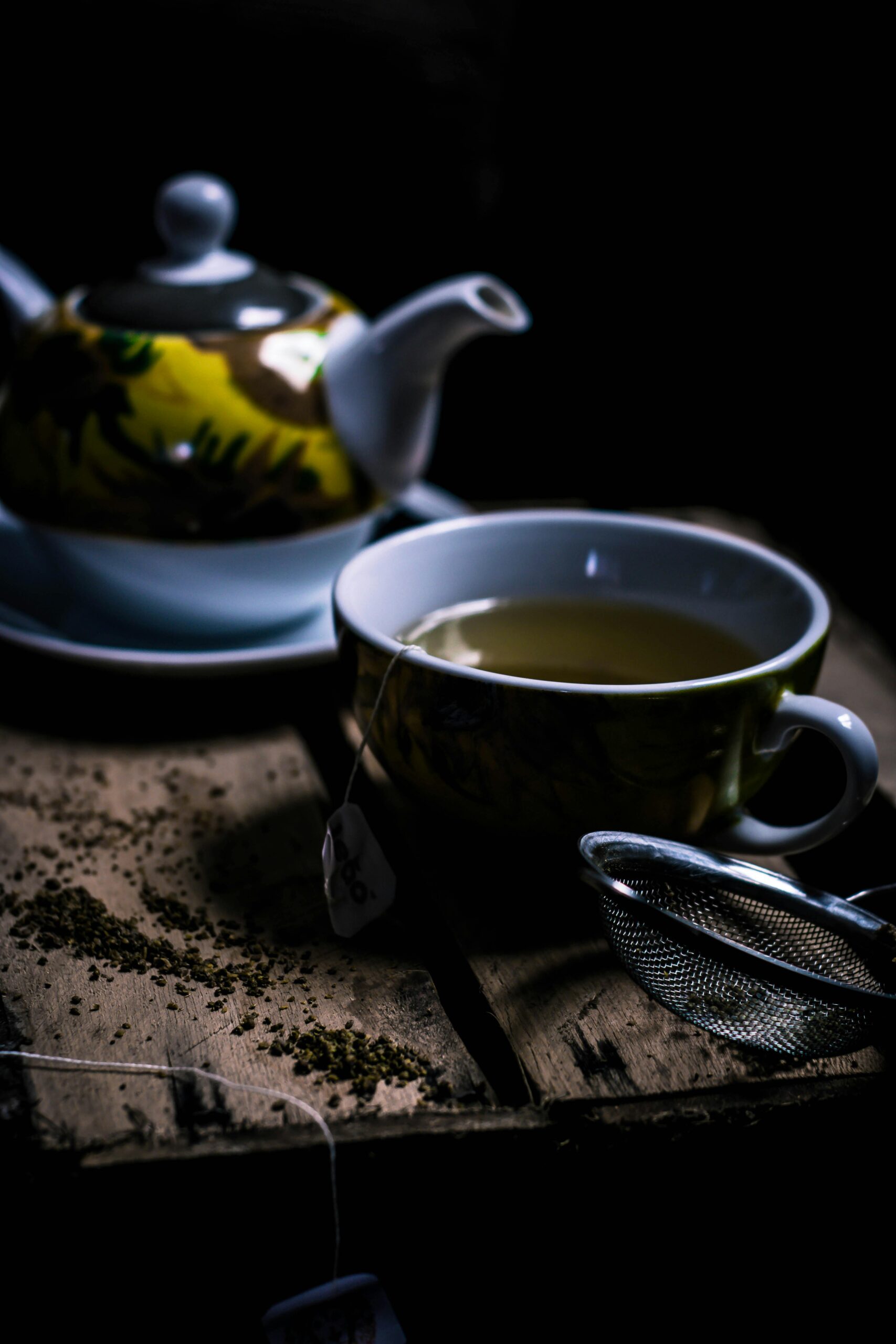 A cozy dark wooden setup with green tea in a teacup, teapot, and strainer.