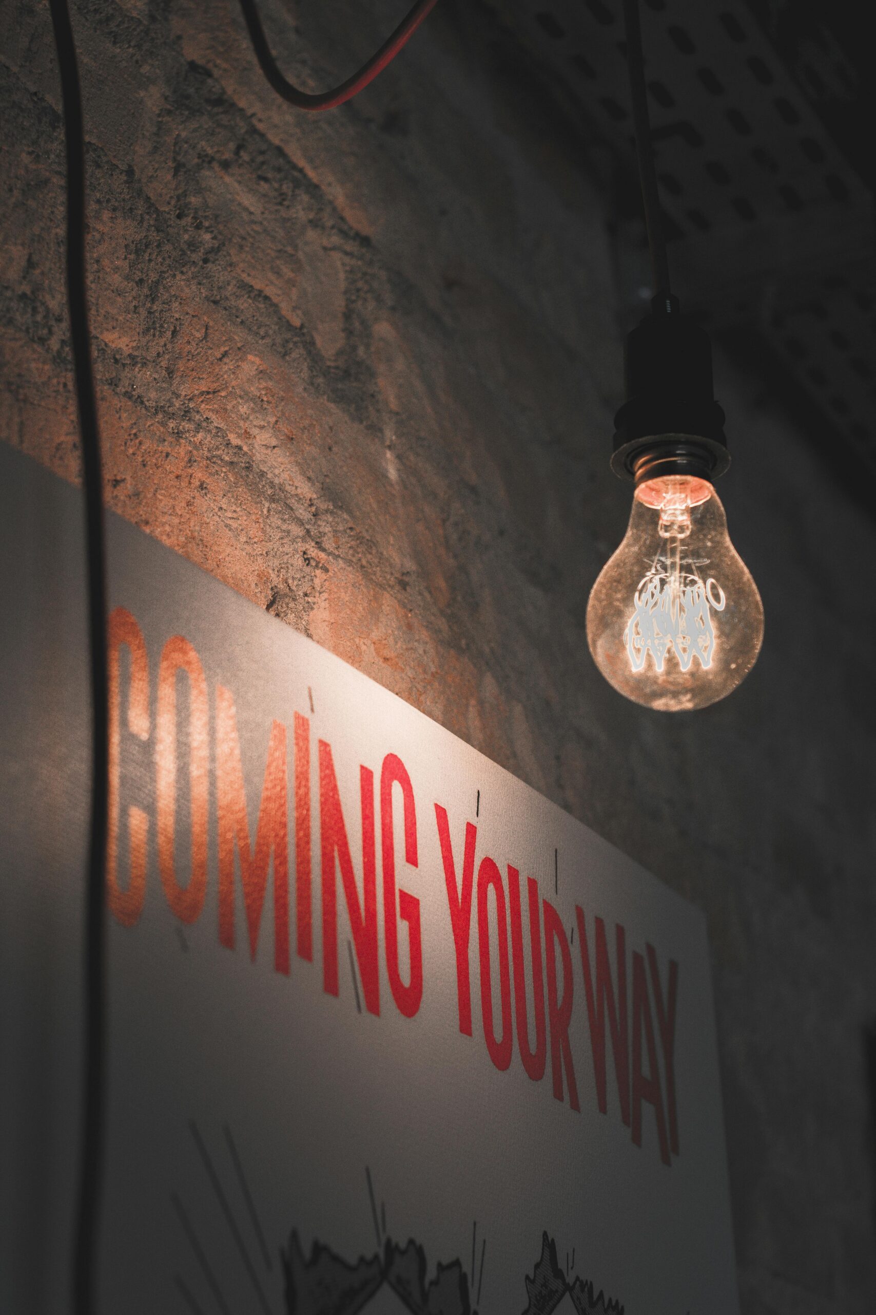 A dimly lit vintage bulb hanging against a rustic brick wall with 'COMING YOUR WAY' sign, evoking urban vibes.